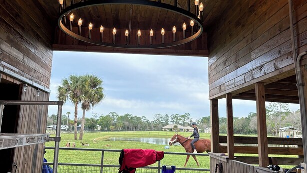 A wooden barn with a circular chandelier, a horse in the background, and a person riding a horse.