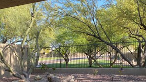 An outdoor patio with a wicker chair, a paved walkway, and a view of a park with trees and a fence. - Anthem Family getaway (Anthem)
