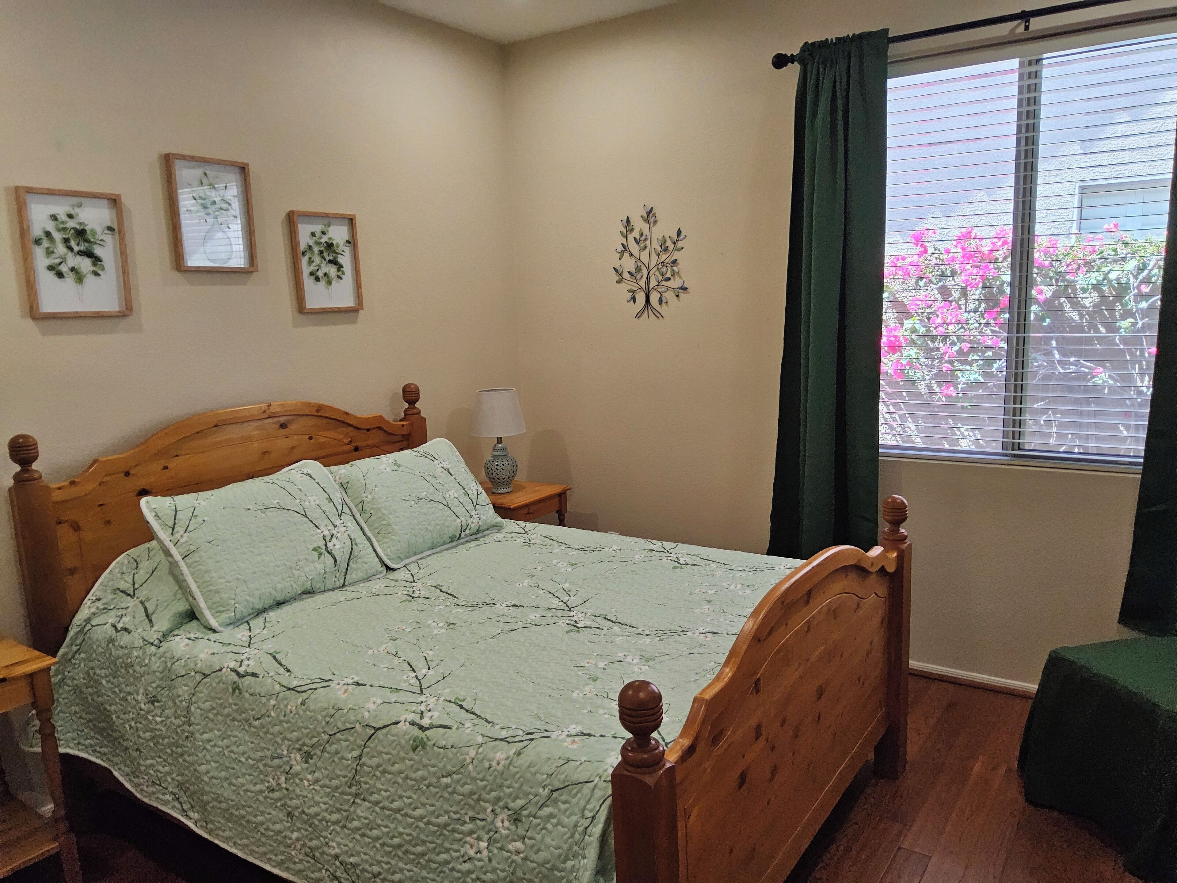 A bedroom with a wooden bed, a bedside table with a lamp, a window with curtains, and three framed pictures on the wall.