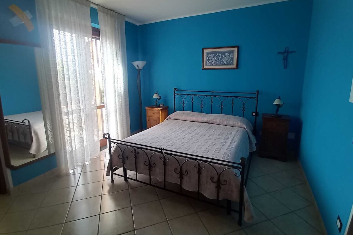 Un dormitorio con cama, dos mesitas de noche, una ventana con cortinas y un cuadro colgado en la pared.