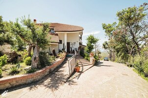Una casa de dos pisos con tejado de tejas, una escalera de piedra que conduce a la entrada, y un jardín con diversas plantas y árboles.