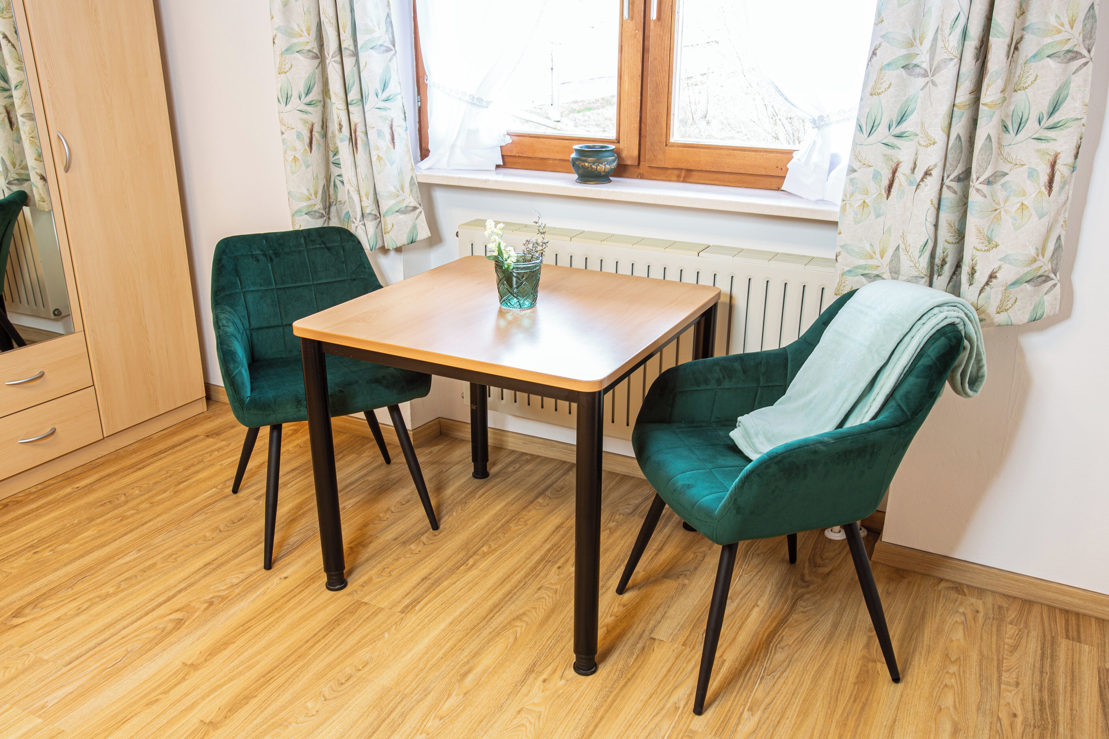 Ein Zimmer mit Holztisch, zwei grünen Stühlen, einem Heizkörper und einem Fenster mit Vorhängen.