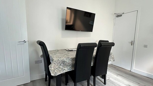 A dining area with a table and four chairs, a mounted flat-screen TV, and a white door. - Carmen Sylva Holiday let Llandudno sea front (Llandudno)