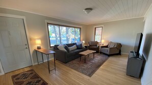 Amplia sala de estar con un sofá gris, una mesa de centro y un mueble para la televisión. Dos ventanas ofrecen luz natural y vistas al exterior.