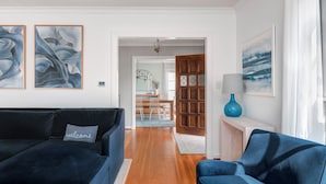 Un salón moderno con un sofá azul oscuro, un sillón a juego y una mesa de comedor de madera con sillas. Hay pinturas abstractas en las paredes y un jarrón azul sobre una mesita auxiliar.