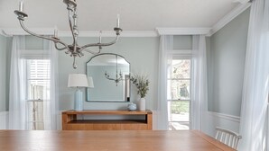 Un comedor con una mesa de madera, sillas blancas, una araña, un espejo y un jarrón con flores.