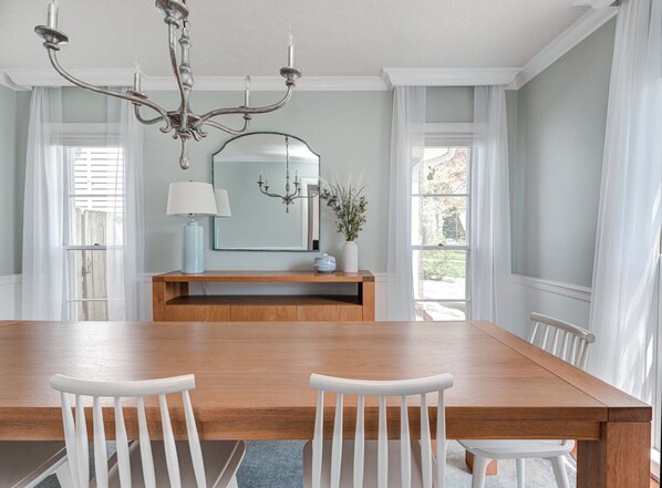 A dining room with a wooden table, white chairs, a chandelier, a mirror, and a vase with flowers.