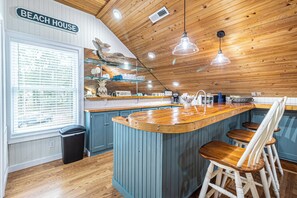 Een keuken met houten kastjes, een houten keukeneiland met krukken, en een bordje met de tekst 'BEACH HOUSE'.