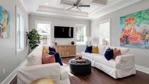 Ein Wohnzimmer mit zwei weißen Sofas, einem schwarzen Hocker, einem Deckenventilator, einem Flachbildfernseher und einem großen Gemälde an der Wand.