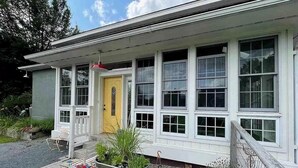 A single-story house with a yellow front door, white trim, and multiple windows. There is a small porch with a bench and potted plants.