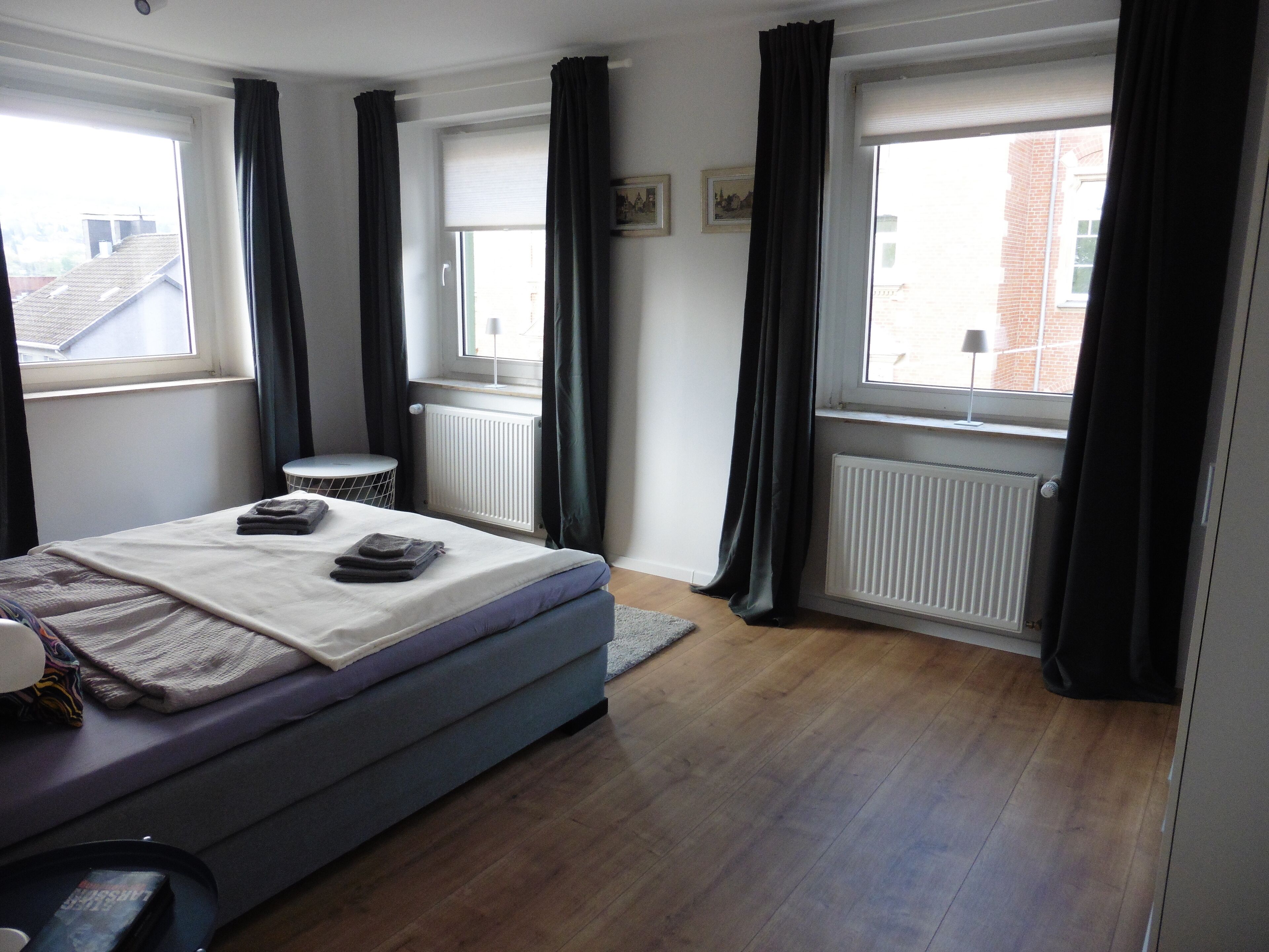 Ein Schlafzimmer mit einem Bett, zwei Fenstern, einer Heizung und einem kleinen Tisch.