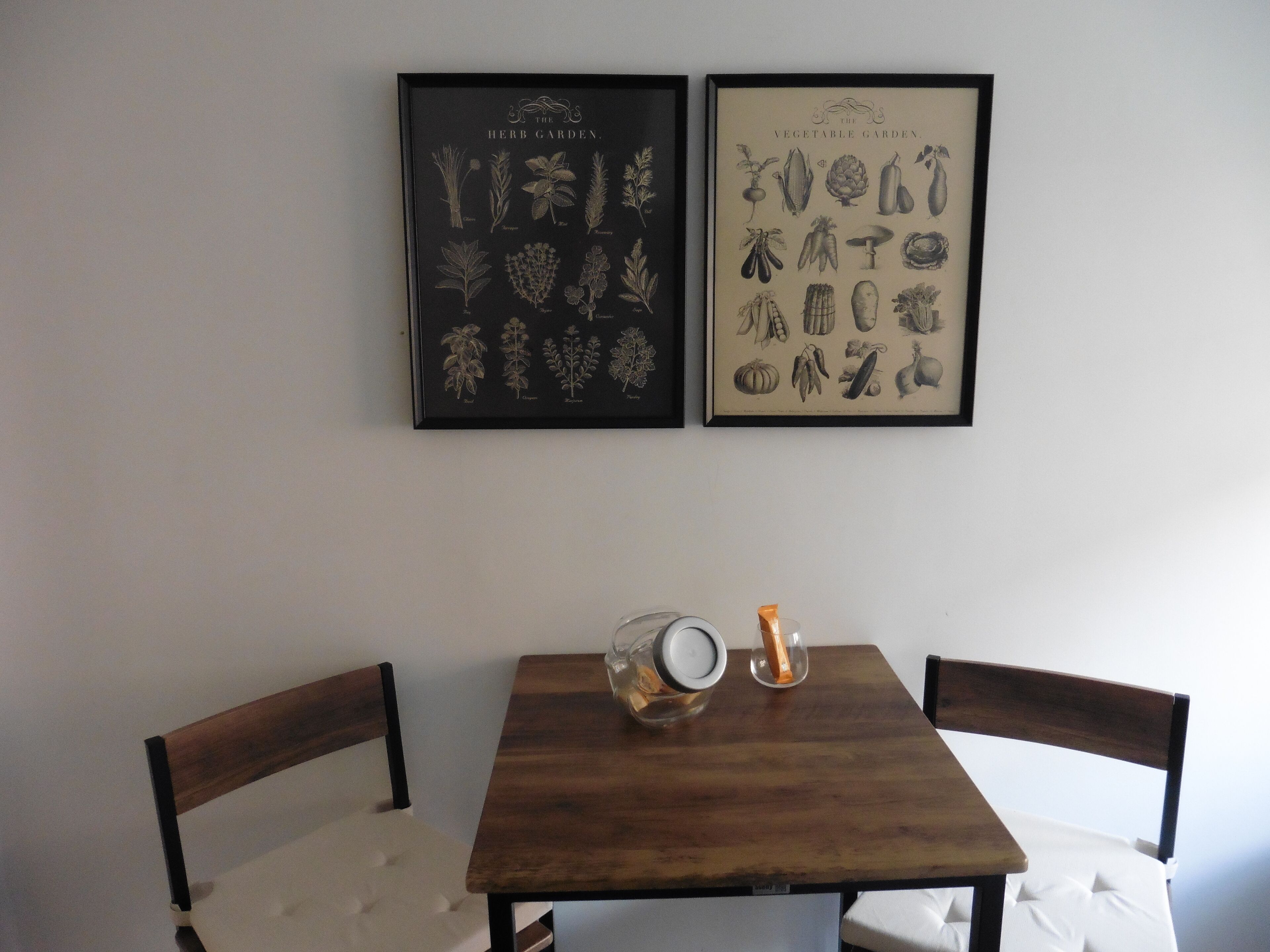 A dining table with two chairs, a framed herb garden, and a framed vegetable garden on the wall.