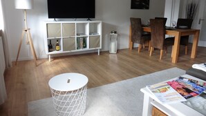 A modern living room with a flat-screen TV, a wooden dining table with chairs, a white cabinet with shelves, a standing lamp, and a magazine rack. - Sedan16 Zentral (Wuppertal)