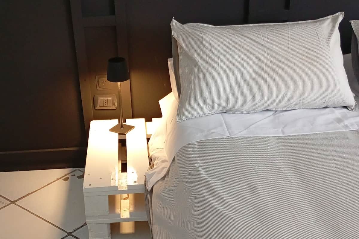A neatly made bed with white linens and a striped pillow, a bedside table with a lamp, and a tiled floor.
