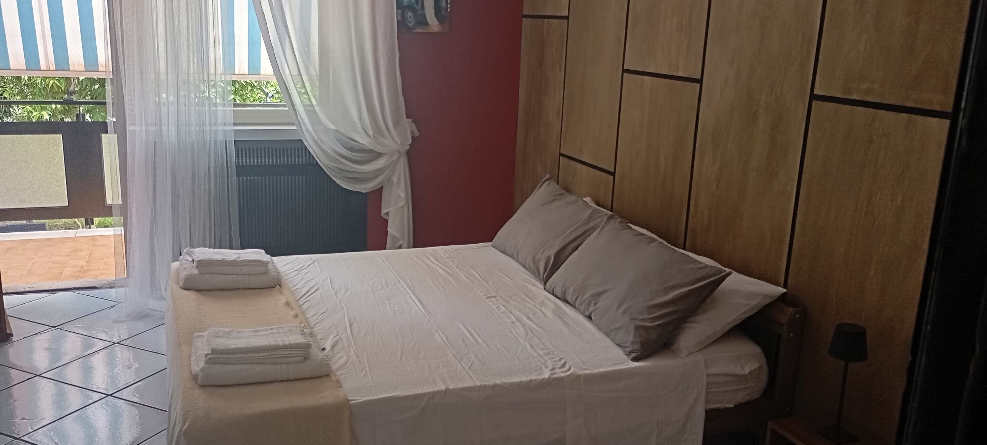 A neatly made bed with white linens and pillows, a wooden headboard, and a window with sheer curtains.