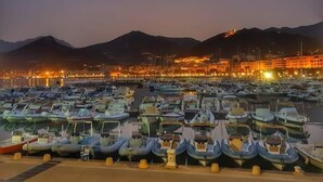 Una marina al crepuscolo con numerose imbarcazioni ormeggiate, una montagna sullo sfondo e un edificio ben illuminato.