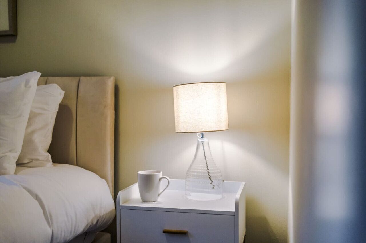 Una cama bien hecha con sábanas blancas, una mesita de noche con una lámpara y una taza.