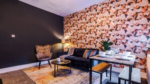 A modern living room with a dark wall, a floral wallpaper, a leather sofa, a round coffee table, and a dining table set with white dishes and glasses. - Salford Sanctuary 5 Bed House (Salford)