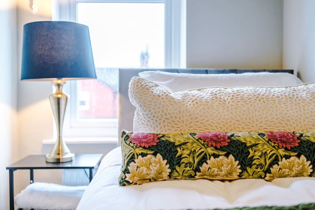 Un lit bien fait, avec un oreiller blanc à motifs, une lampe bleu foncé posée sur une petite table à côté, et une fenêtre en arrière-plan.
