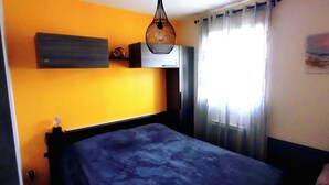 A room with a bed, a hanging light fixture, a wall-mounted shelf, and a window with curtains. - Proche Mer, Appartement Classé 2 Dans Petite Résidence (Santa-Lucia-di-Moriani)