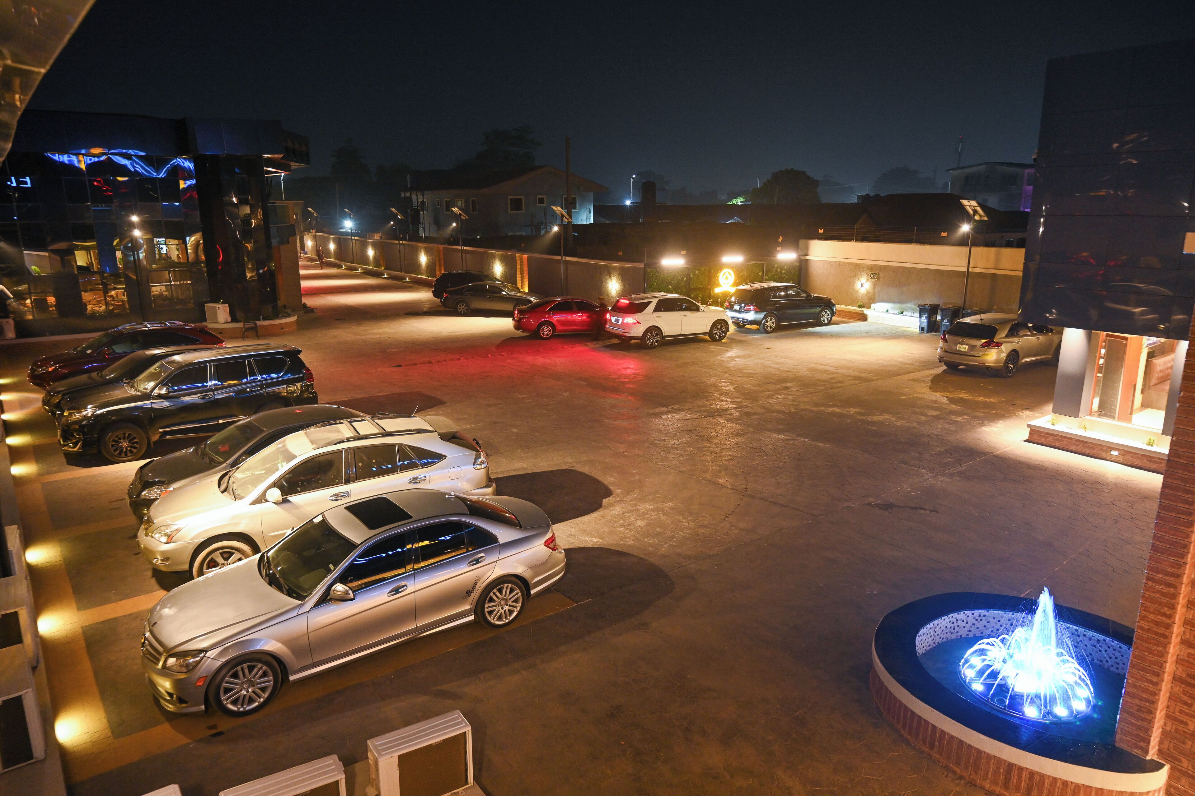 A parking lot with multiple cars, a fountain, and a building with illuminated signage.