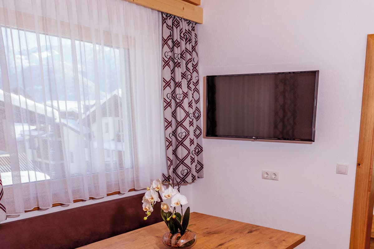 A room with a wooden ceiling, a TV mounted on the wall, a wooden table, a sofa, and a window with sheer curtains.