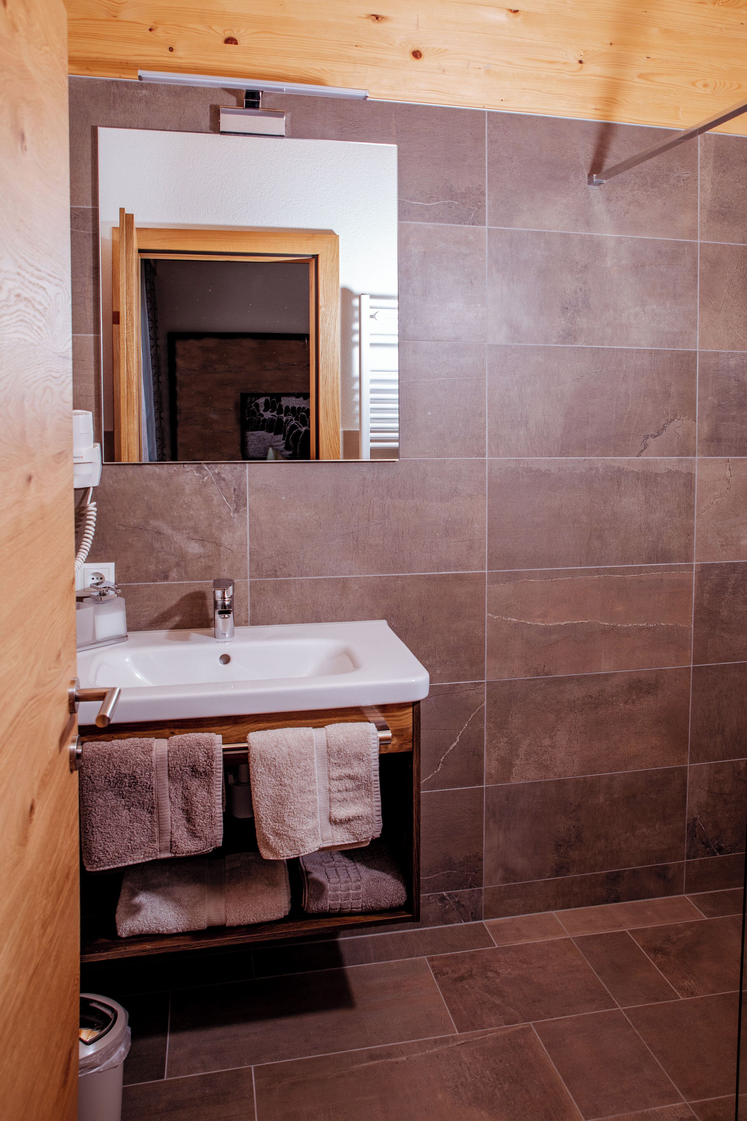 A bathroom with a wooden ceiling, a white sink, a mirror, and brown tiles.