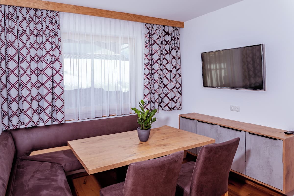A modern living room with a wooden dining table, cushioned seating, a potted plant, and a flat-screen TV mounted on the wall.