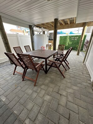 Un área de patio techada con una mesa y sillas de madera, una cerca blanca y un seto verde al fondo.