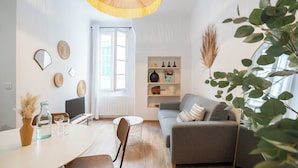 A modern living room with a sofa, dining table, and chairs. There is a window with blinds, a hanging light fixture, and a shelf with decorative items. - T2 Tout Confort - Central- Bord de mer 100m - Clim (La Ciotat)