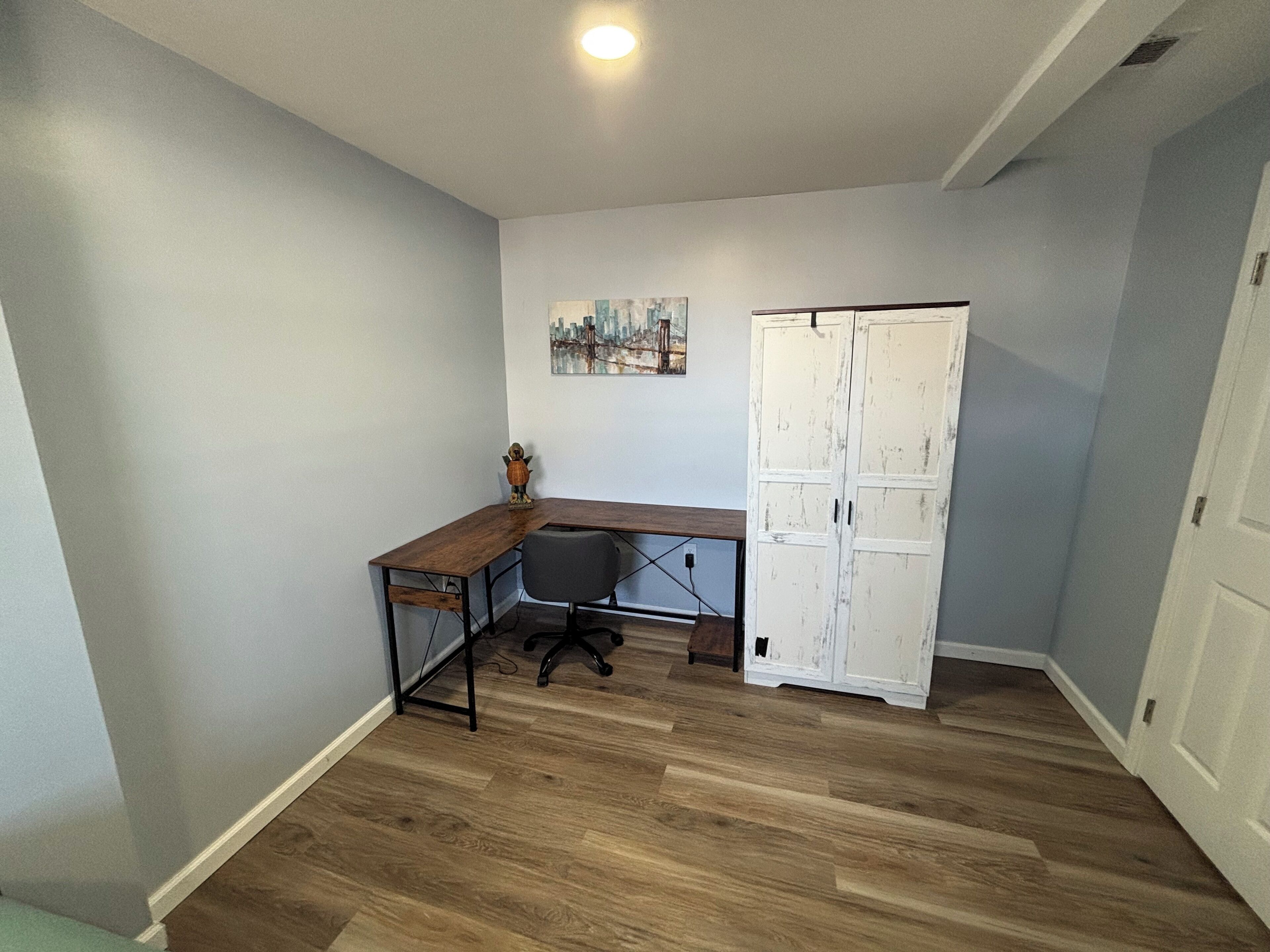 Een kamer met een houten bureau, een stoel, een witte kast en een stadsgezichtschilderij aan de muur.