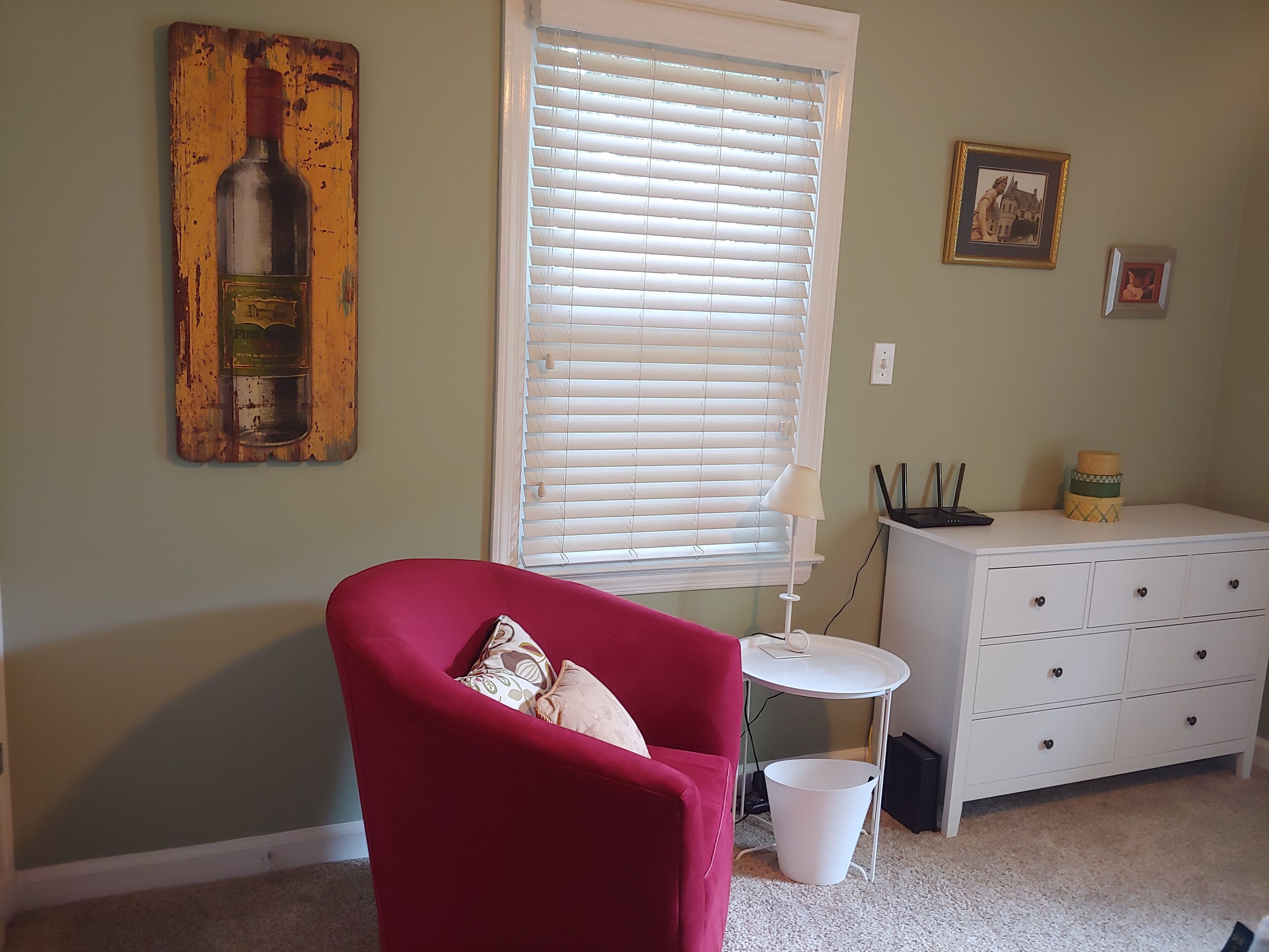 Une pièce avec un fauteuil rouge, une commode blanche, une fenêtre avec des stores, une lampe et un tableau représentant une bouteille.