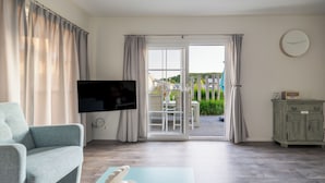 A living room with a sofa, a TV, a table with a decorative item, a cabinet, and a sliding glass door leading to an outdoor area with a table and chairs. - Luxuriöses Chalet in Fußnähe zum Strand (Biggekerke)