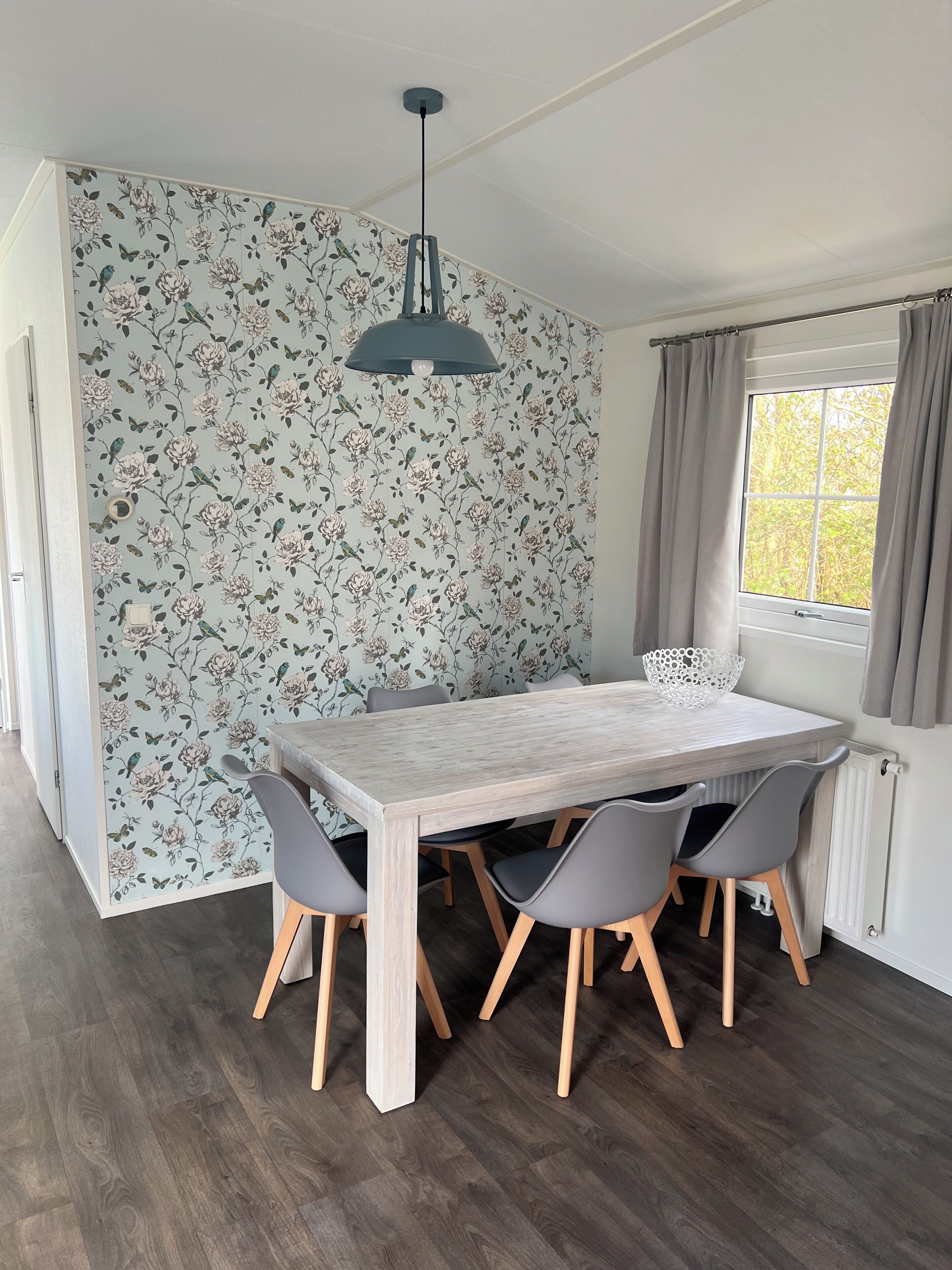 Un coin repas agrémenté d’un papier peint à motifs floraux, d’une table en bois, de chaises grises et d’un luminaire suspendu.