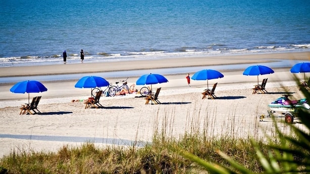 A beach with several blue umbrellas and beach chairs, a bicycle, and a person walking. - Luxury 6-Bedroom Home with Private Pool, Elevator & Family-Friendly Comfort (Hilton Head)