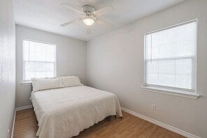 A bedroom with a bed, two windows, a ceiling fan, and hardwood flooring.