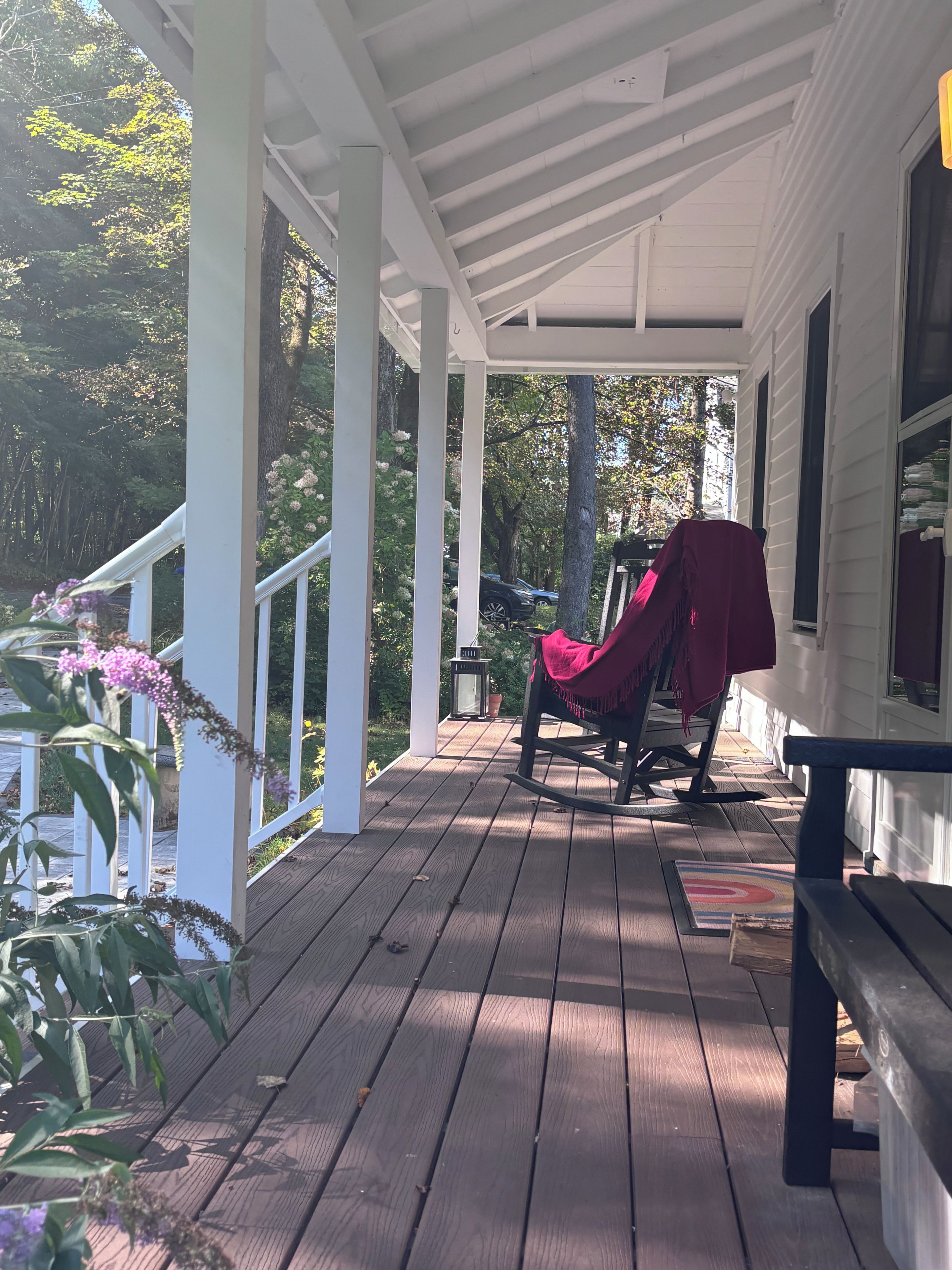 A wooden porch with a rocking chair and a bench, surrounded by greenery and trees.