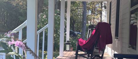 A wooden porch with a rocking chair and a bench, surrounded by greenery and trees.