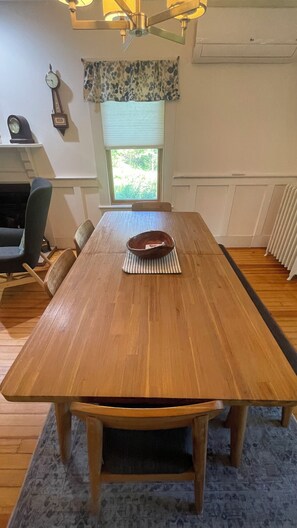 Une table à manger en bois, avec des chaises, une horloge et une fenêtre avec des stores.