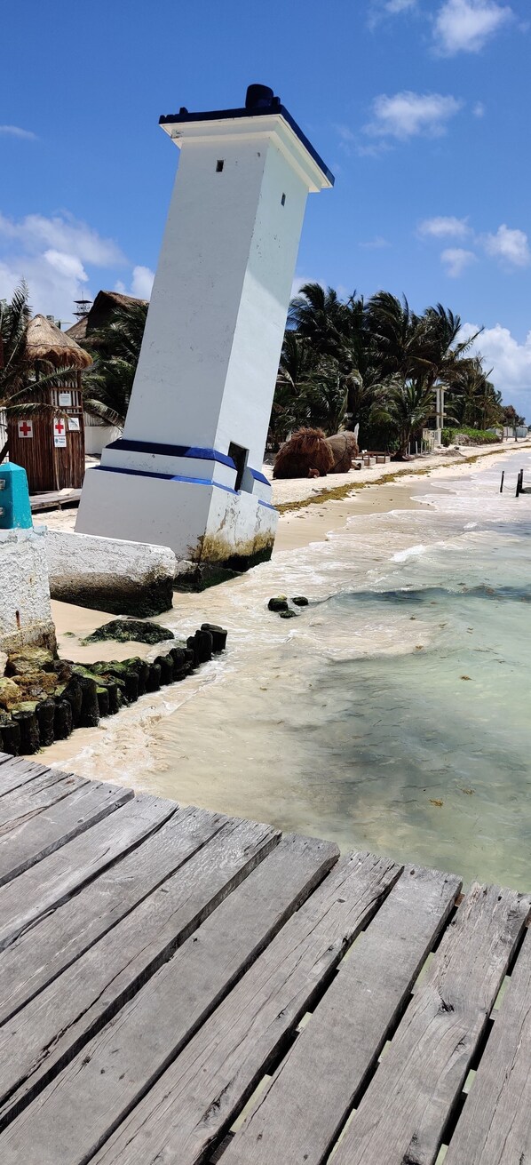 A wooden pier leading to a white lighthouse with a blue stripe, surrounded by a sandy beach and palm trees.