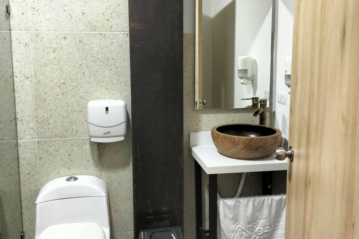 A bathroom with a toilet, a trash bin, a soap dispenser, a wooden sink, and a mirror.