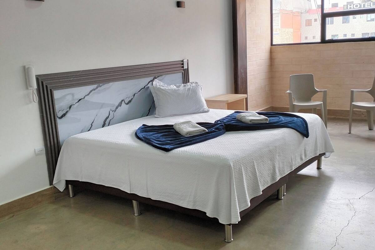 A hotel room with a large bed, a small table, two chairs, and a window with a view of buildings.