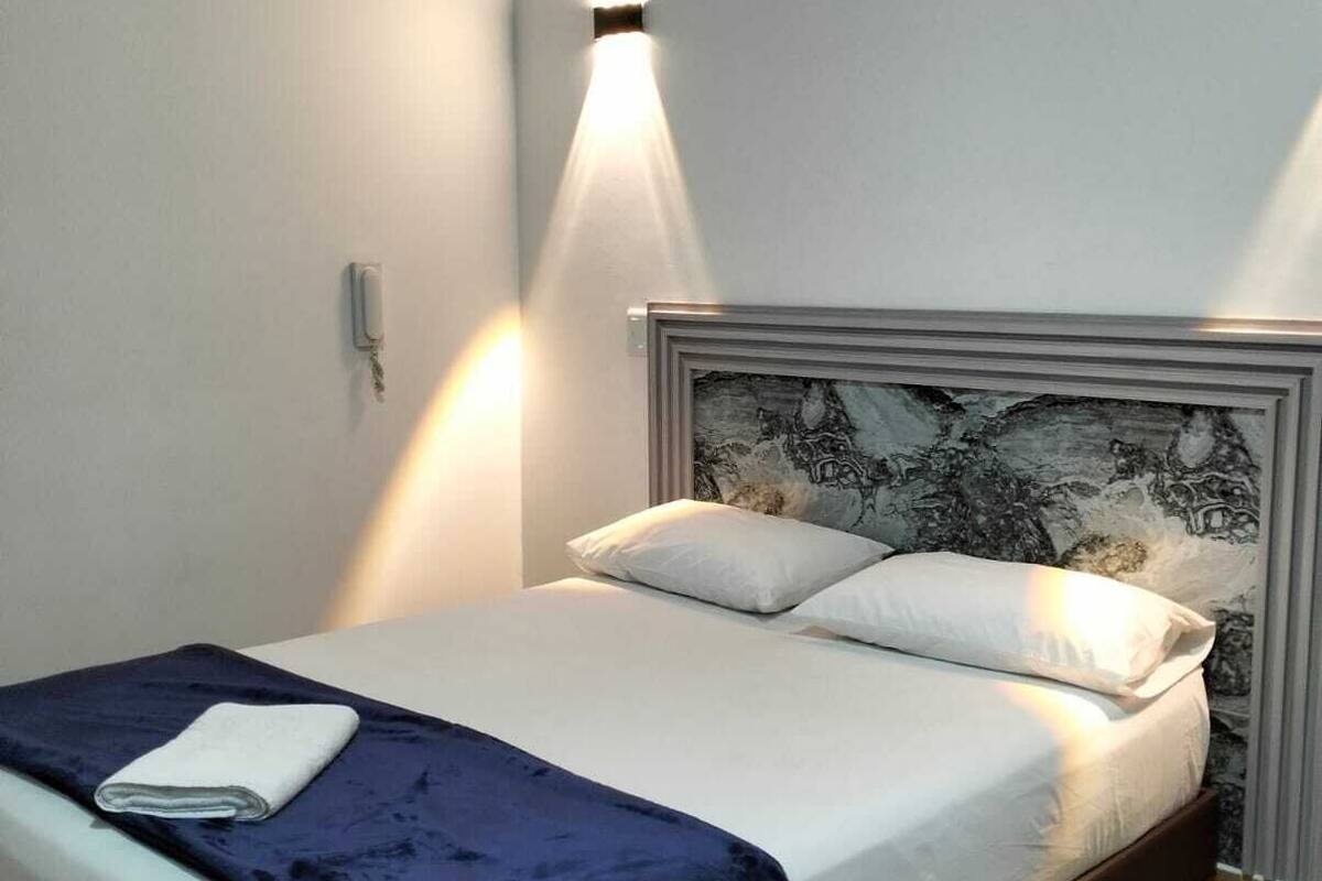 A neatly made bed with a dark blue blanket and white linens, a headboard with a patterned design, and a wall-mounted light fixture.
