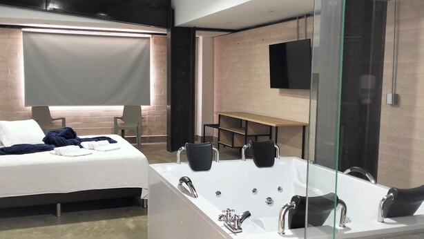 Habitación de hotel moderna con una cama grande, un área de estar separada, una bañera espaciosa con múltiples grifos, un box de ducha de vidrio y un televisor de pantalla plana montado en la pared.