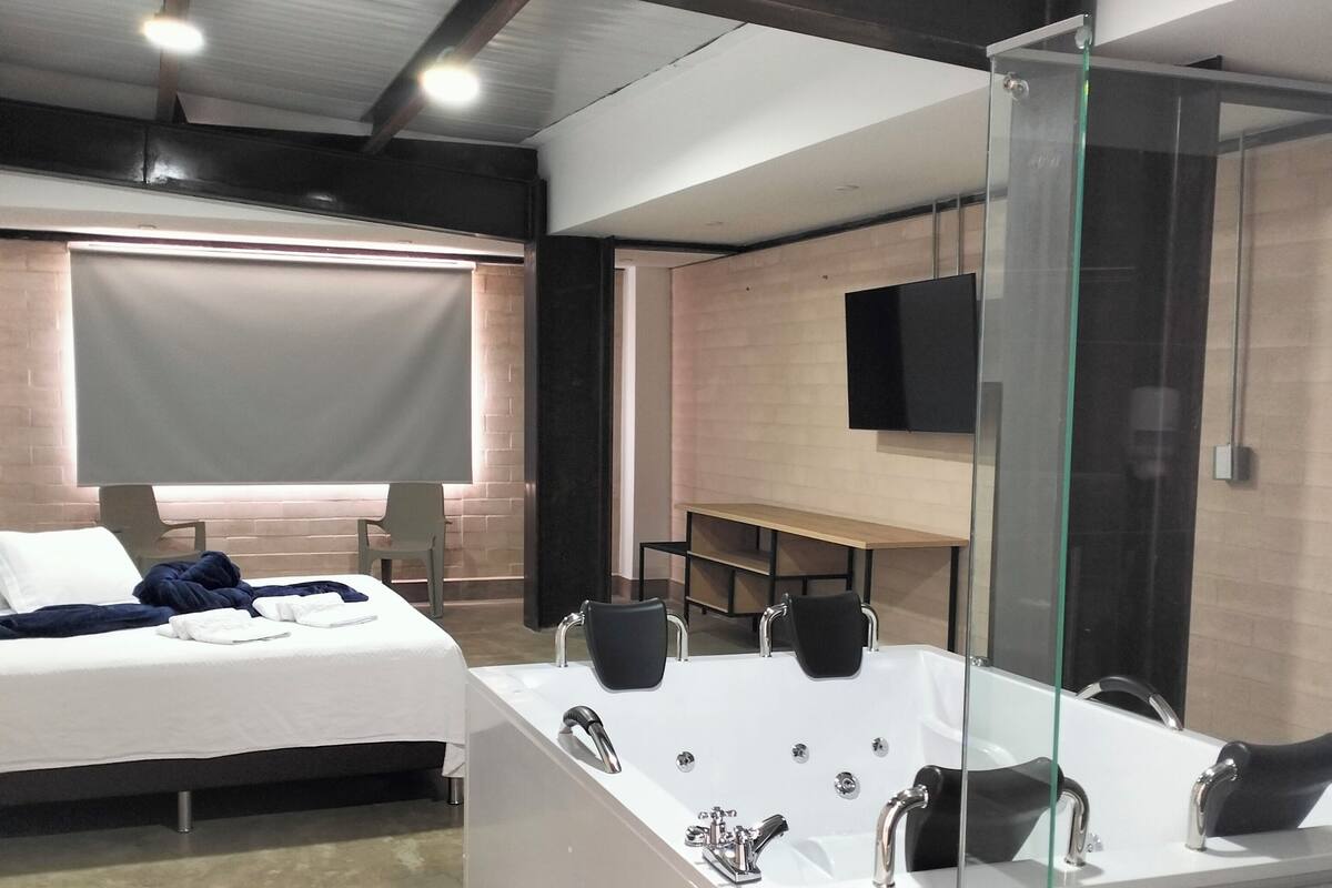 A modern hotel room with a large bed, a separate sitting area, a large bathtub with multiple faucets, a glass shower enclosure, and a flat-screen TV mounted on the wall.