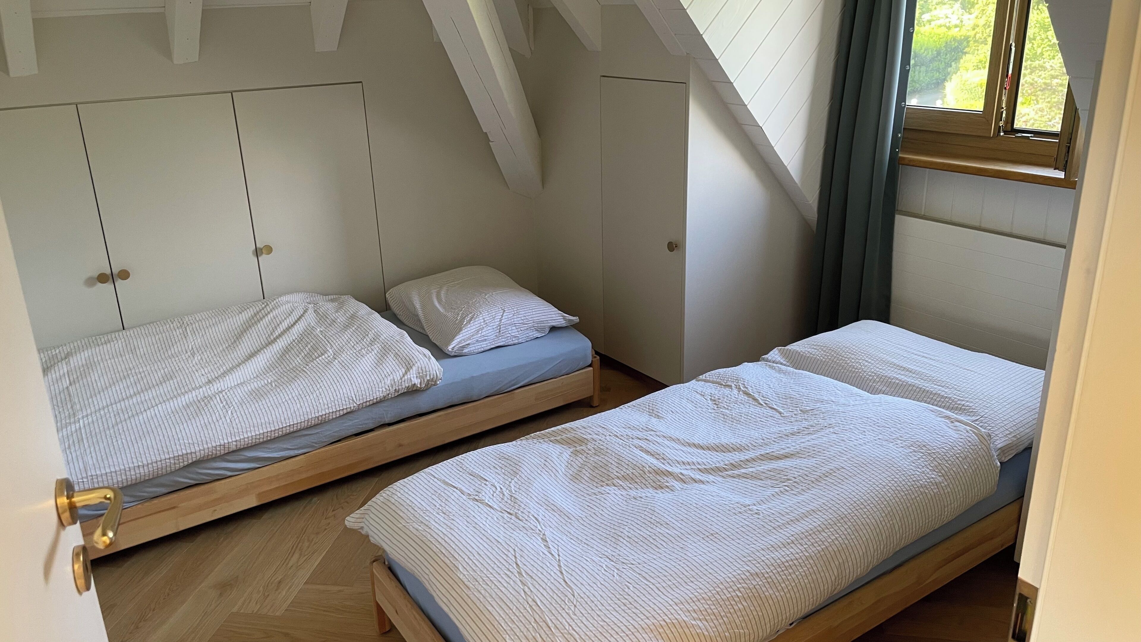 Twee eenpersoonsbedden met houten frames en gestreept beddengoed in een kamer met witte wanden en een schuine balkenkap.