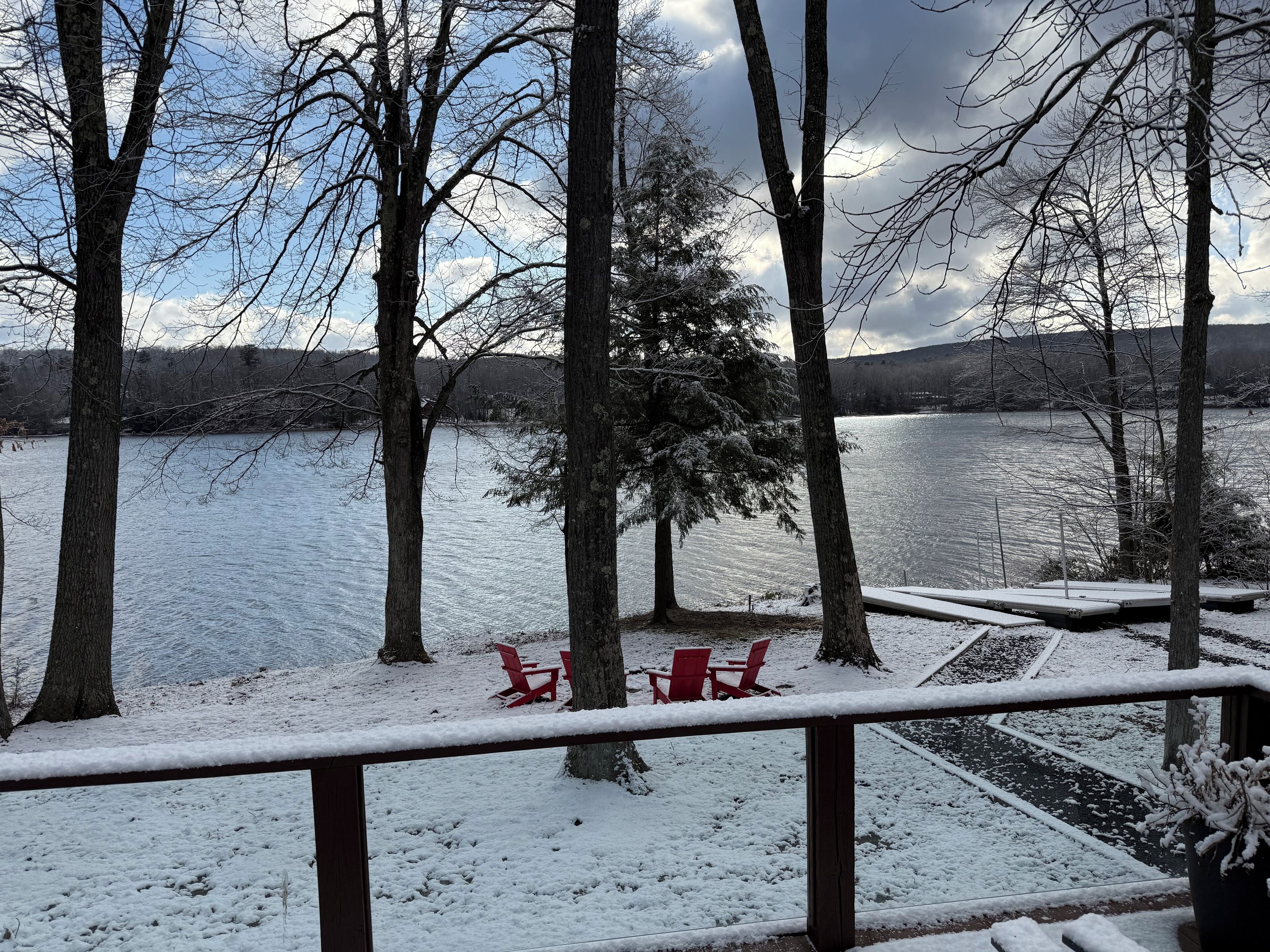 A snowy deck with red chairs and a view of a lake and trees.