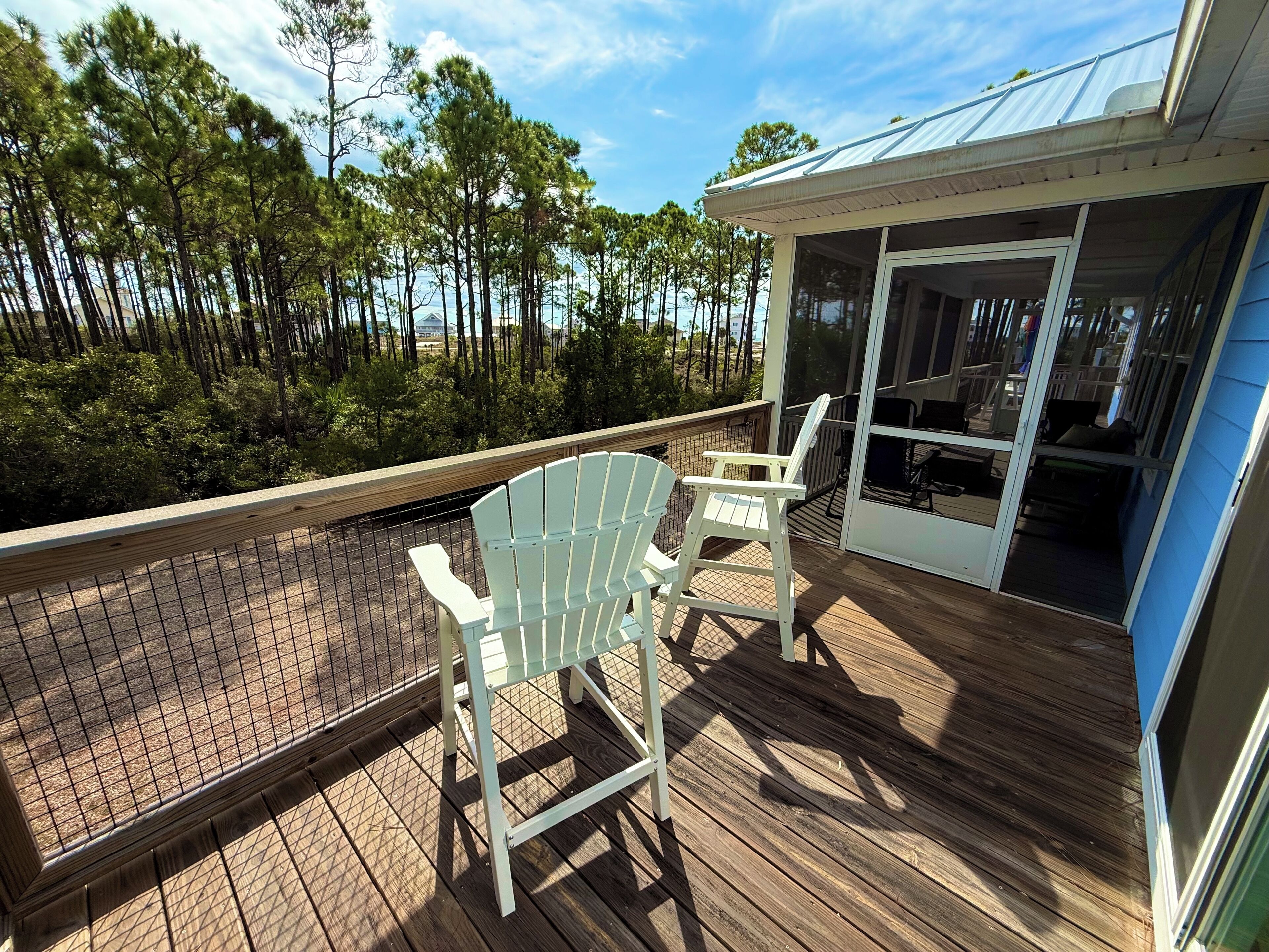 Une terrasse en bois avec deux fauteuils Adirondack blancs, une porte vitrée donnant sur un espace intérieur et une vue sur une zone boisée.
