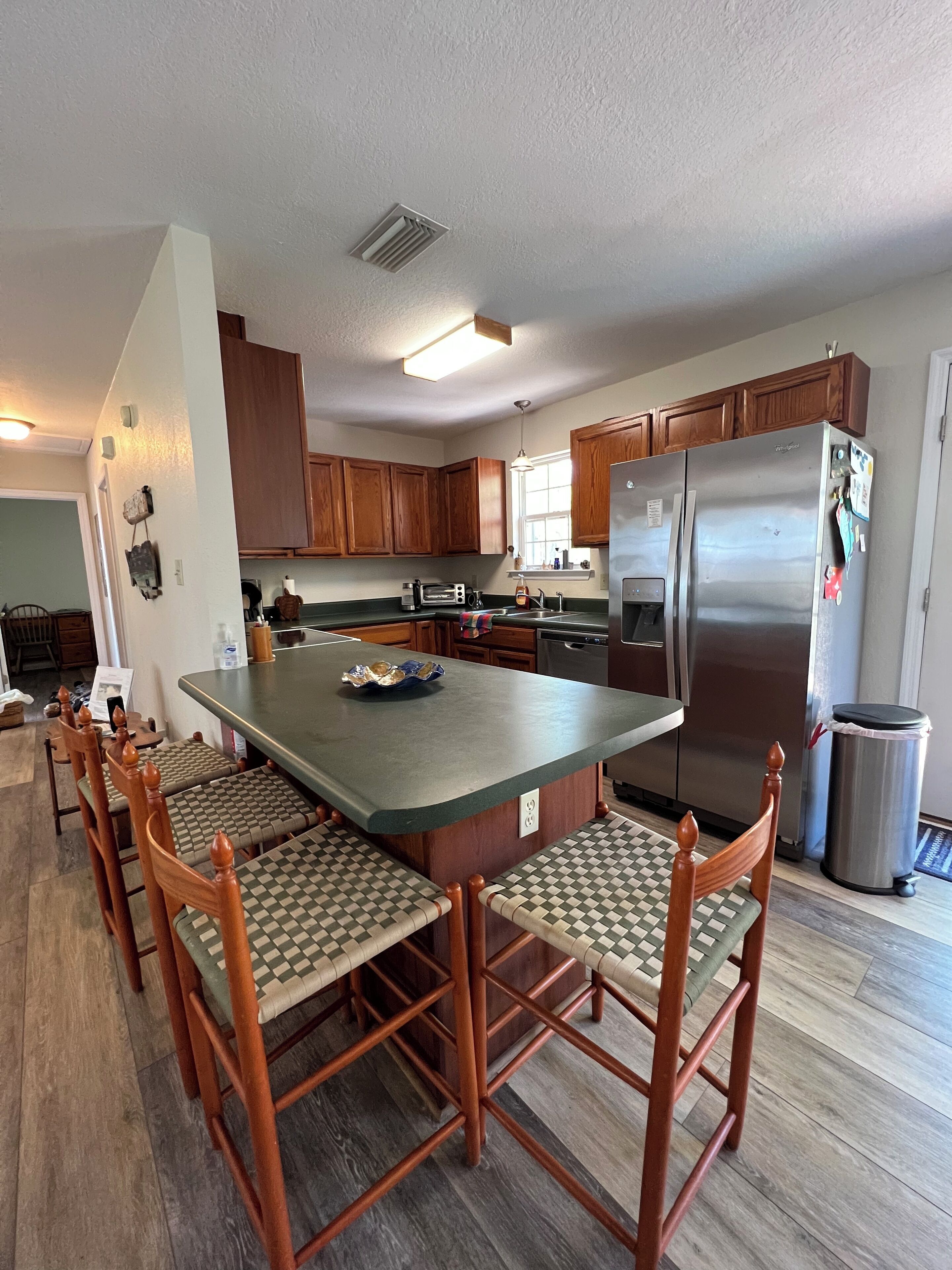 A modern kitchen with a large island, wooden chairs, a stainless steel refrigerator, and wooden cabinets.