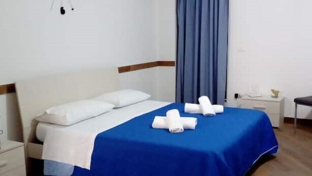 Un llit ben arreglat amb un estovat blau i almoades blanques, una taula de nit amb una làmpada i una cortina blava.